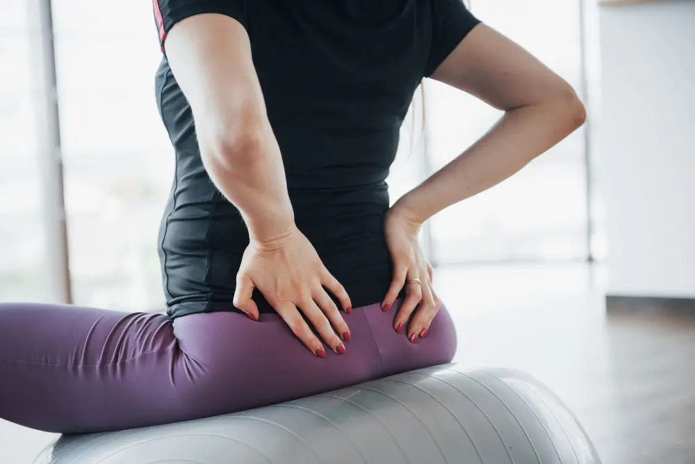 Woman holding lower back and hip area while seated on exercise ball, representing piriformis syndrome and sciatica pain